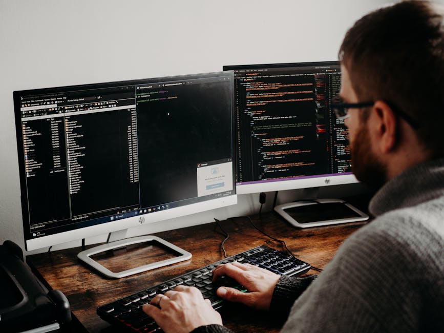A man with a beard and glasses working at a wooden desk with dual computer monitors displaying lines of code and software interfaces. The workspace includes a black keyboard, a mouse, and a smartphone