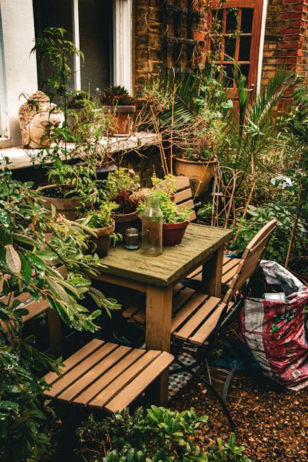 A cozy outdoor garden area adjacent to a brick house, featuring a weathered wooden dining table surrounded by matching wooden chairs with slatted seats. The table is topped with various potted plants,