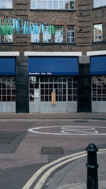 The exterior of the Shoreditch Arts Club building features a brick facade with several large windows, some with white frames. Above the entrance, a blue awning displays the venue's name. On the street